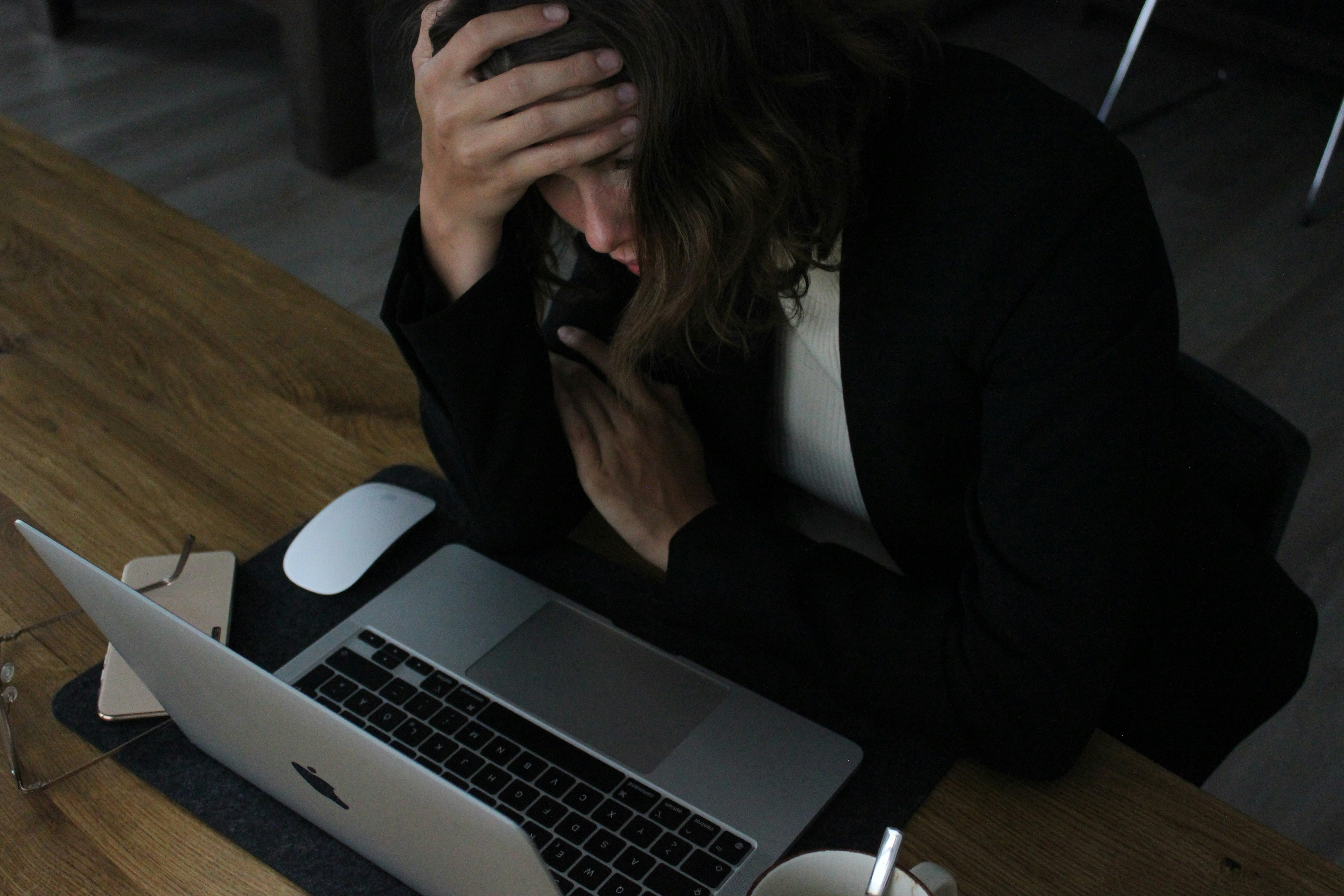 Stressed professional woman with hands on head sitting at desk surrounded by papers and laptop showing overwhelm and burnout
